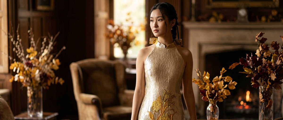 A woman stands in a warmly lit, vintage-style room wearing a sleeveless cream cheongsam / qipao dress with gold embroidery and gold geometric earrings. The room features a fireplace, wooden paneling, and dried floral arrangements.